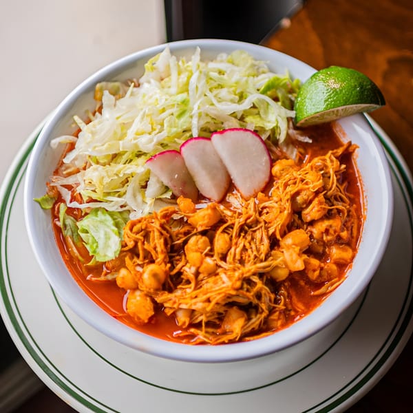 A bowl of El Arriero Taqueria's pozole with shredded chicken, hominy, cabbage, radishes, and lime.