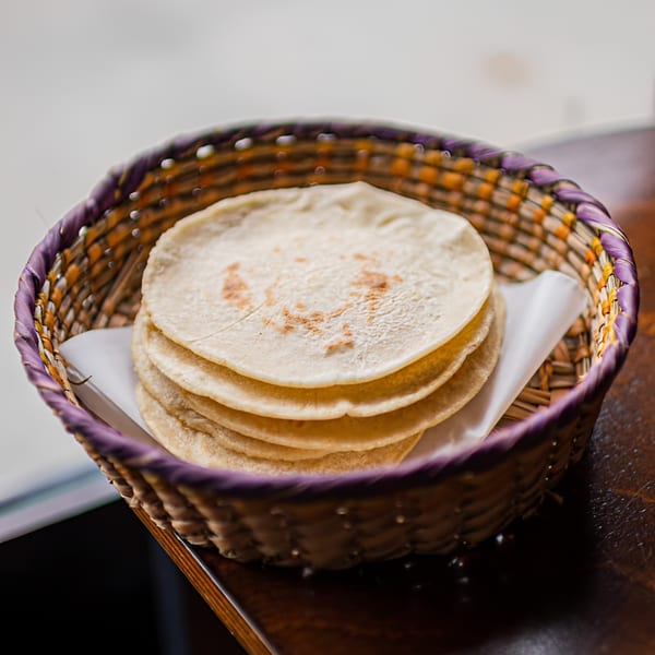 A woven basket with purple and orange trim holds a stack of warm corn tortillas.