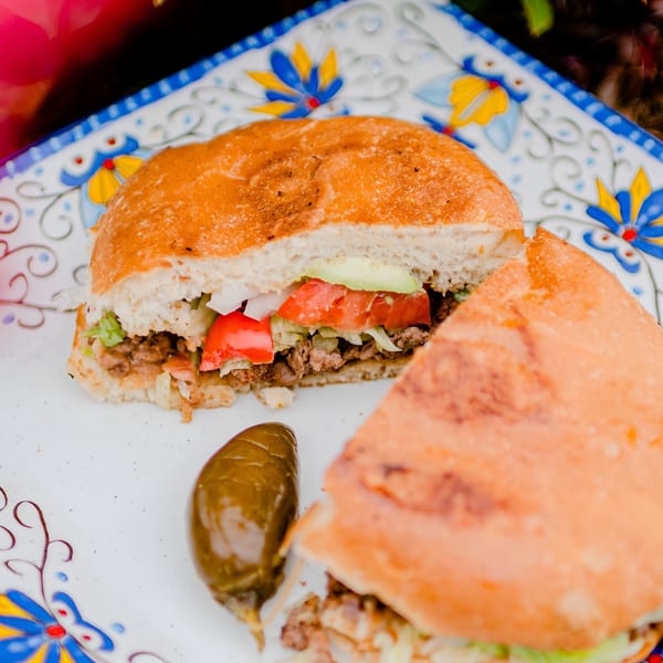 A Mexican torta sandwich, cut in half, with meat, lettuce, tomato, and avocado, served with a pickled jalapeño.