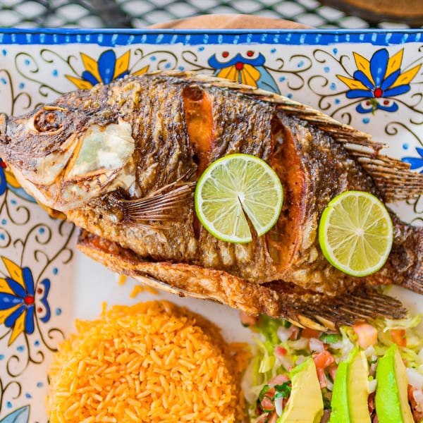 Whole fried fish with lime slices, orange rice, and a side salad with avocado on a decorative plate.
